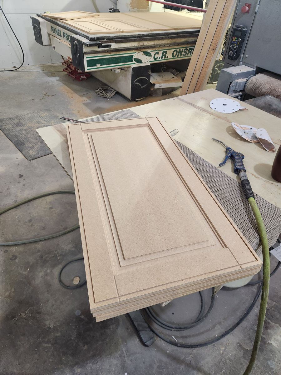 Stack of one-piece raised panel MDF cabinet doors on a shop bench, with the CNC router behind them